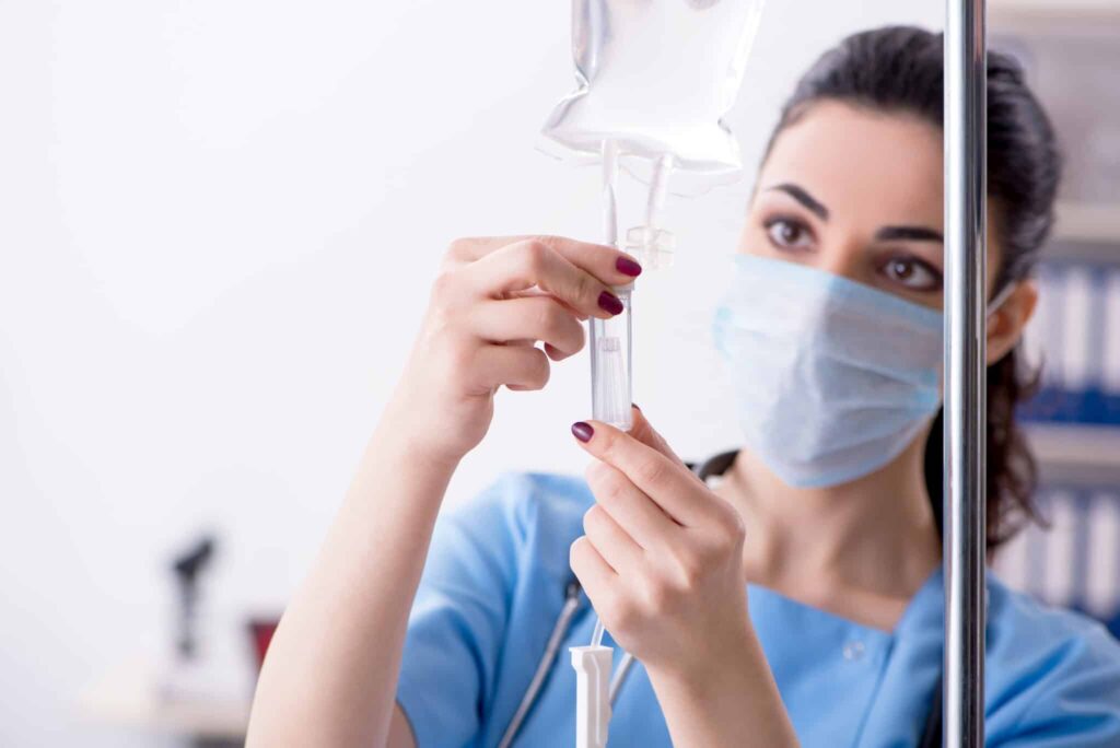 A masked nurse inspecting and preparing an IV fluid bag for use in a hospital environment.