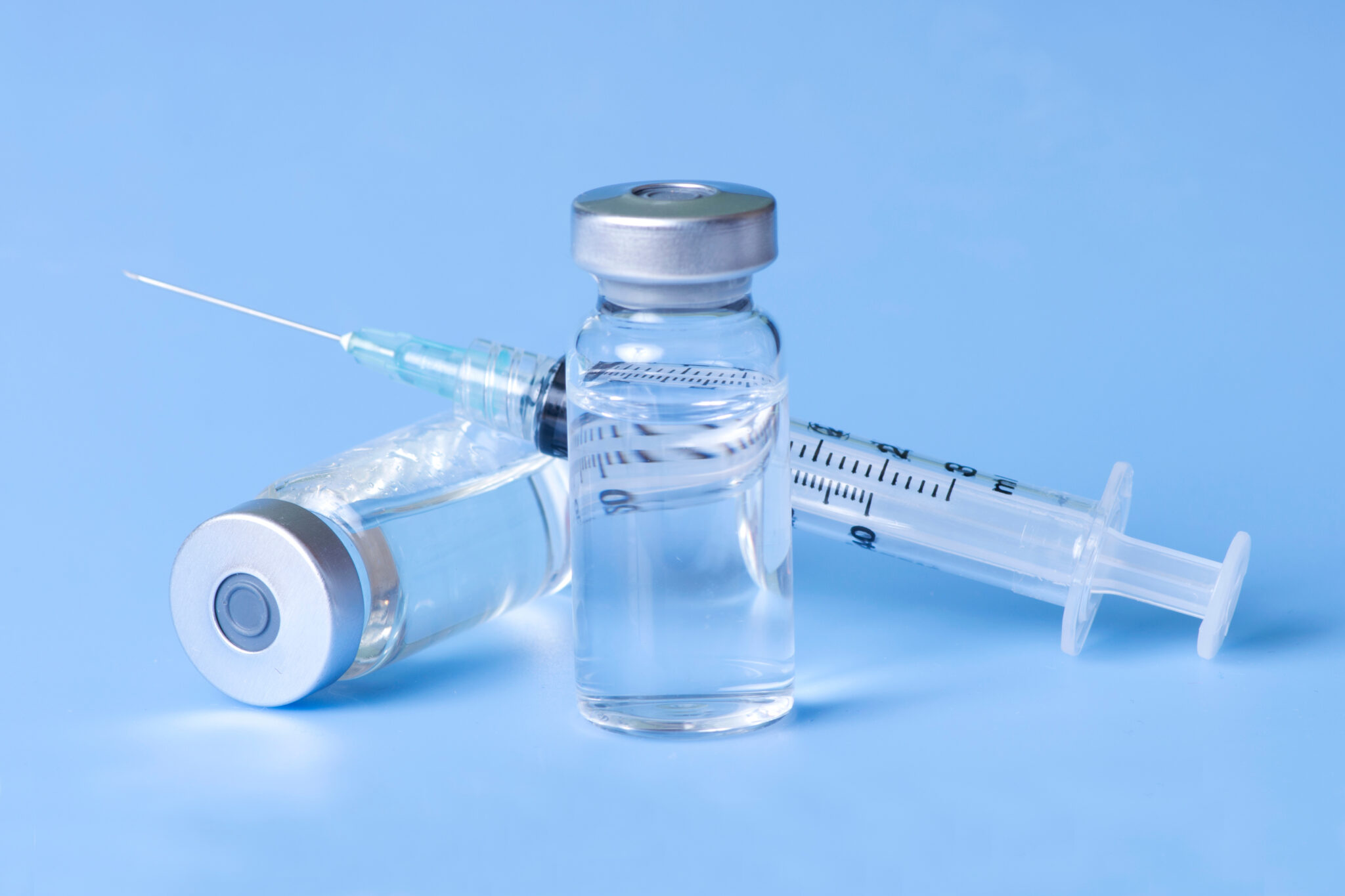 Close-up of a clear medical vial and syringe on a light blue background, filled with a transparent liquid.