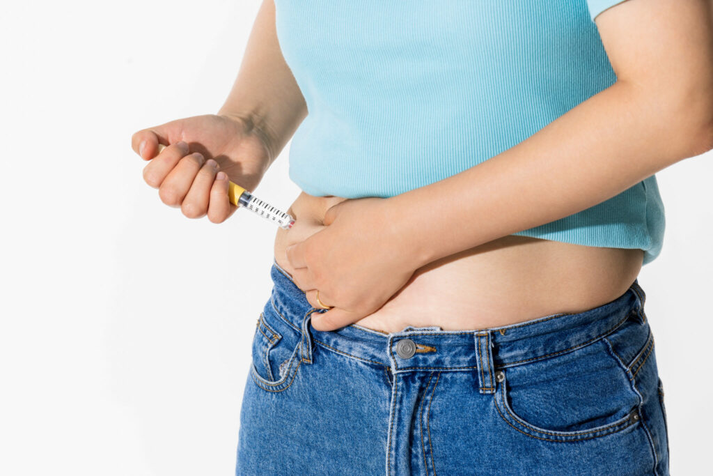 Close-up of a person in a blue shirt and jeans administering a subcutaneous injection into their abdomen using a syringe.